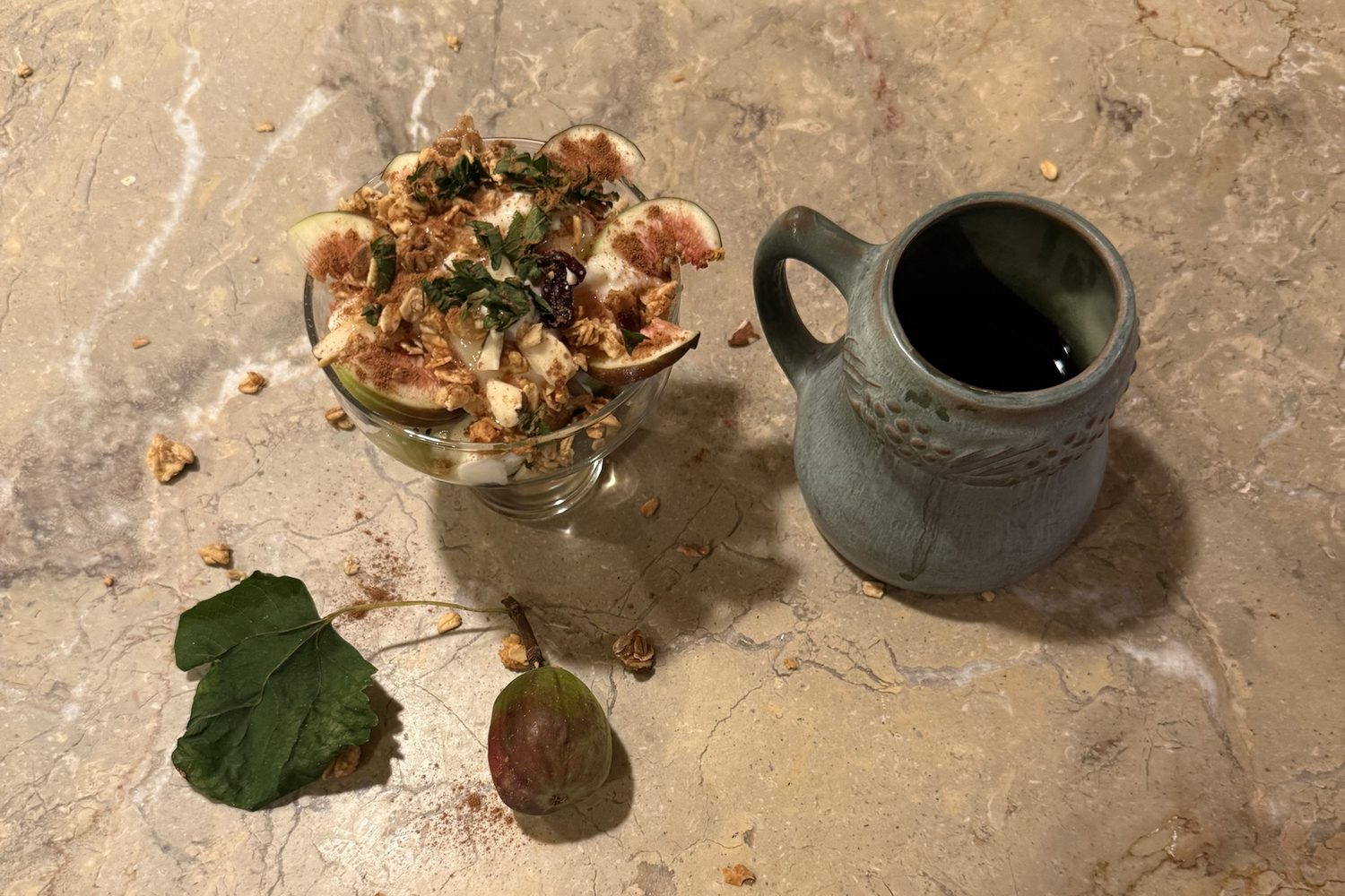 Seen from above on a honey-coloured marble counter: a glass coupe of yogurt layered with sliced fresh figs, granola, almonds and chopped mint, dusted with cinnamon; beside it a green stoneware pitcher, and in front a whole fig with its leaf